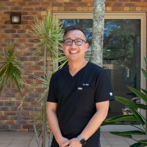 Dr William Luong, Senior Orthodontist at Medland Orthodontics standing in front of a brick wall with windows, smiling beautifully towards the camera