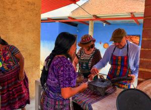 Mayan woman smiling and cooking alongside international travelers in a traditional Guatemalan kitchen, teaching ancestral Mayan recipes.