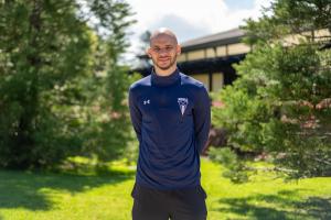 Coach Tj Curry is a man who is standing outdoors in front of a building and surrounded by green trees, wearing a navy blue long-sleeve athletic shirt with the Menlo College logo and Under Armour branding, smiling at the camera with his hands behind his ba