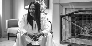 Anne Marie Otáñez seated and smiling in a stylish monochrome portrait, wearing a white suit, with a fireplace and modern decor in the background.