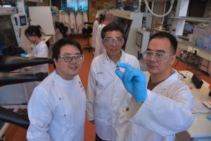 The image shows three male scientists in white lab coats, holding a small lead free perovskite solar cell in the shape of a chip.