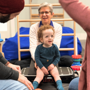 A toddler sits on a balance board, surrounded by two adults and colorful toys, in a playful, supportive environment.