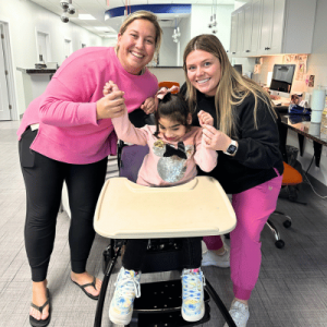 Two women wearing bright sweaters assist a young girl in a high chair, smiling in a brightly lit room with modern decor.