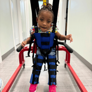 A child wearing a blue supportive harness and pants sits on a rehabilitation apparatus in a bright, clinical setting.