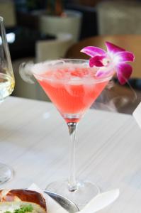 A pink cocktail served in a martini glass with a purple orchid garnish and visible vapor, placed on a white table.