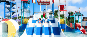 Two children slide down bright blue and white water slides at a vibrant water park, enjoying a sunny day with playful attractions in the background.