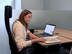 Female researcher sitting at desk reviewing printed materials while working on laptop computer with Transkriptor software, creating accessible educational content for students.