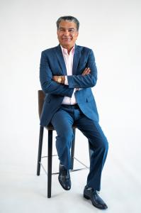Man in a blue suit and white shirt sitting on a tall wooden stool against a white background, looking confidently at the camera.