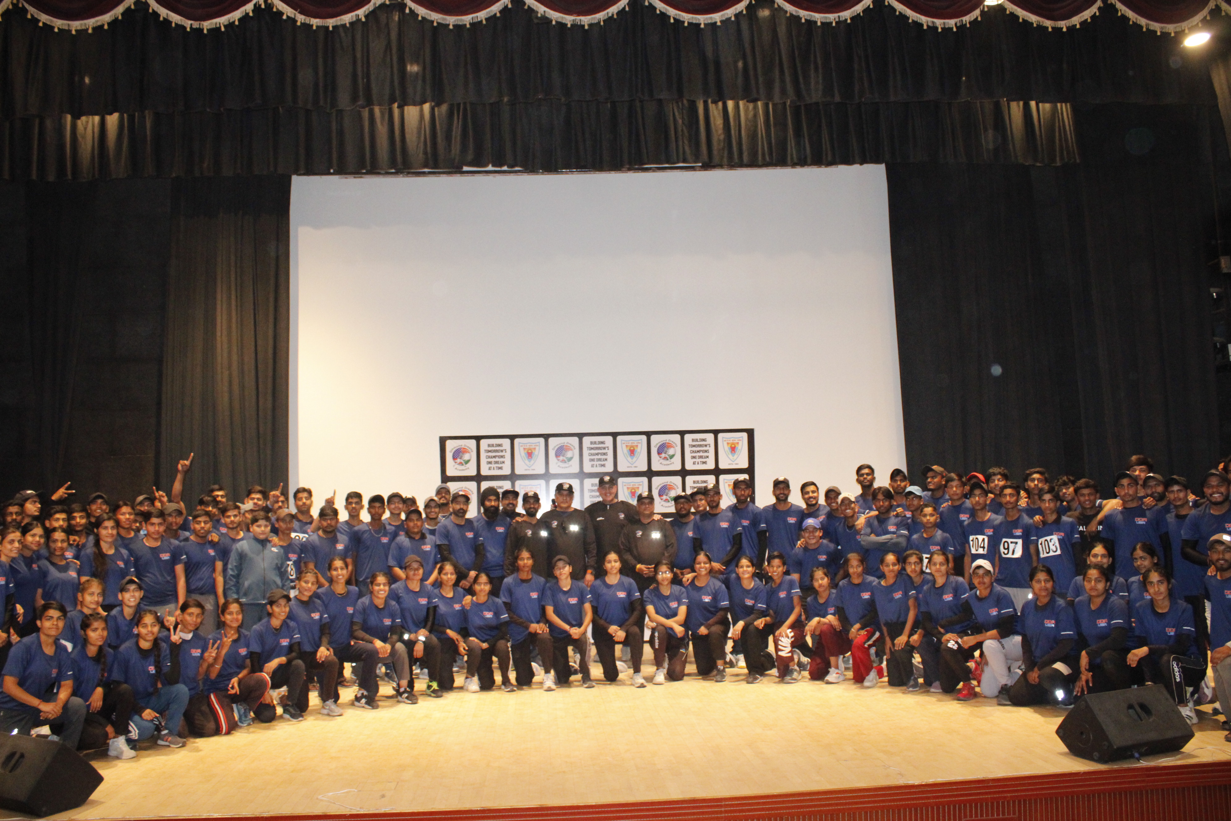 DDA players, coaches, leadership team and staff pose together in Black DDA T-shirts and Blue DDA jerseys for a group photo at a DDA Camp, showcasing their participation and camaraderie.
