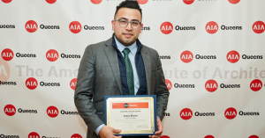 Bravo in a suit holds a certificate while standing in front of a step-and-repeat banner with AIA Queens and The American Institute of Architects logos.