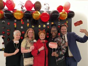 Five adults stand together smiling in front of a Las Vegas-themed photo backdrop decorated with red, black, and gold balloons, playing card symbols, and dollar signs. Each person is holding playful props like bowties, signs, and fake cards. The group is d
