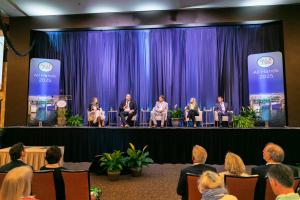 A group of five panelists, seated on stage, speak to a large crowd of attendees.