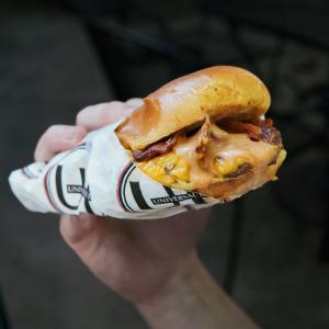 A close-up of Universal Joint’s smash burger with melted cheese, crispy bacon, and sauce, held in a hand and wrapped in UJ-branded paper. This is the free burger guests receive on National Burger Day, May 28, by joining the Universal Joint Loyalty Program