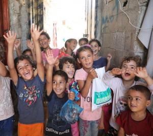 Group of smiling and cheering children in a narrow alley, with one child holding a Qurbani meat bag from Al Mustafa Welfare Trust.