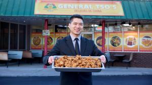 Patrick Cheng standing in front of General Tso’s restaurant holding a large tray of fried chicken, highlighting the business’s catering success.