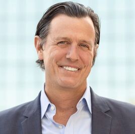 Smiling man with short dark hair wearing a navy blazer and light blue shirt, standing in front of a blurred glass building background.