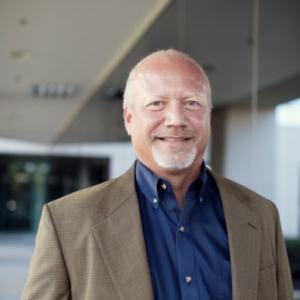 Smiling man with a light beard and short white hair, wearing a brown blazer over a dark blue shirt, standing outside near a modern office building with glass walls.
