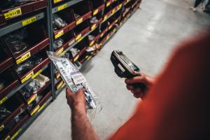 A person scans a packaged HEMI engine part in a well-organized automotive parts warehouse, with labeled bins for Mopar components.