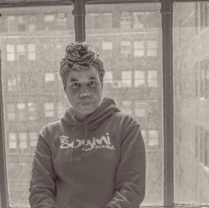Ayana Soyini standing in front of a large open window leaning elegantly against a radiator wearing a Soyini Sound hoodie and an African headwrap.
