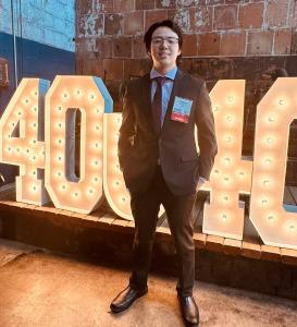 Max Kopp wearing a dark suit and tie, standing in front of large lighted numbers “40 Under 40” at the Globe Dye Works venue in Philadelphia.