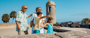 A family stands on a stone fort with cannons and palm trees in the background, enjoying a sunny day together.