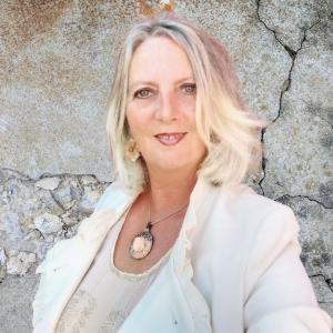 Portrait of Swanette Kuntze, a visionary strategist and founder of Green & Glam, standing against a textured stone wall, wearing a white blazer and pendant necklace.