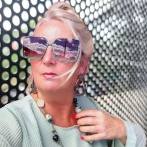 Close-up portrait of Swanette Kuntze wearing oversized sunglasses reflecting clouds and sky, standing in front of a perforated metal wall, styled in pastel green with bold jewelry.
