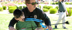 A police officer guides a child in archery at an outdoor event, surrounded by greenery and participants in the background.