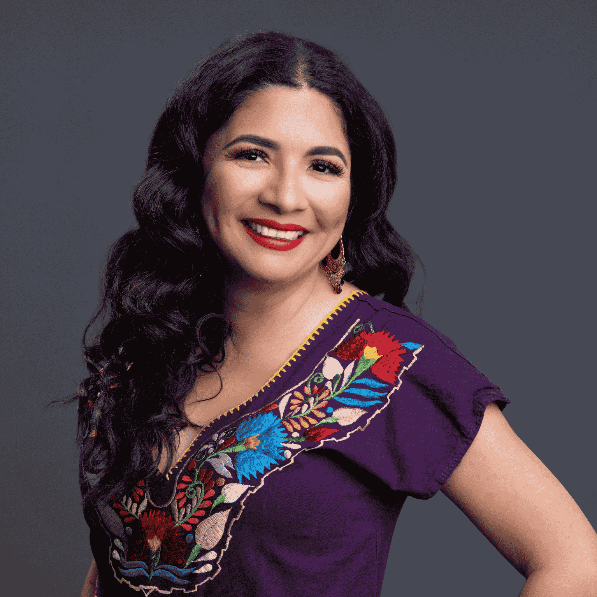 Sandra Guzman smiling in front of a dark gray background, wearing a purple blouse with colorful traditional Mexican embroidery, representing her cultural pride and heritage.