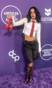 17-year-old rock artist Jaexlynne stands confidently at the 2025 American Music Awards, holding a striking pair of red heels, dressed in punk-inspired fashion, embodying the spirit of rock rebellion.