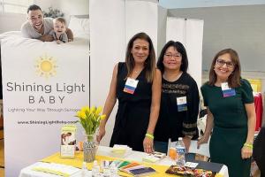 Three women—Mariana Martinez, Vicky Li, and Zara Griswold—stand behind a booth for Shining Light Baby, smiling at an event with informational materials on display.