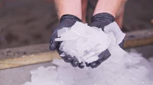 Hands in black gloves hold a pile of jagged ice shards, set against a blurred background of more ice. The scene conveys coldness and precision.