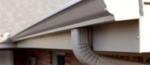 Close-up of a clean beige rain gutter on a house with a downspout, set against a blurred brick wall.