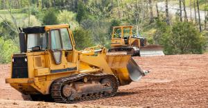 Two yellow bulldozers operate on a freshly cleared dirt site surrounded by lush green trees, conveying an active construction scene.