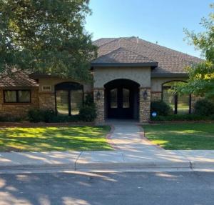 Single-story house with stone facade, arched doorway, and large front windows, surrounded by a well-maintained lawn and trees, under a clear sky.