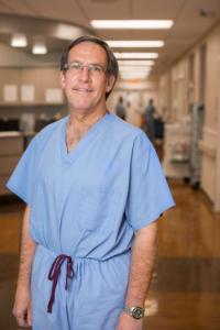 John A. Griswold, M.D., wearing light blue scrubs, stands in a clinical facility hallway  