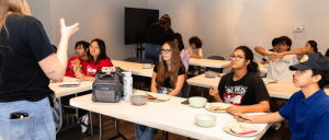A group of people seated at tables in a classroom setting, engaged in conversation and sharing a meal, with plates and bowls in front of them.