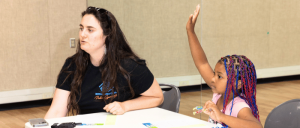 A young girl with colorful braided hair raises her hand while sitting at a table, engaging in a discussion or activity.