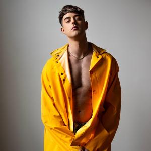 Colin Grafton, a young athletic man wears a bright yellow raincoat which is unbuttoned, revealing his shirtless torso. He poses for the camera, promoting the film "Nosebleed".