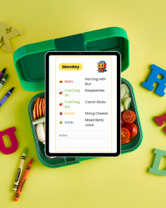 A green lunchbox filled with food items sits beneath a tablet displaying a Monday lunch packing card from the Lunchbox Hero app, showing selected foods like hot dog, raspberries, carrots, string cheese, and raspberry lemonade.