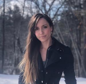 Woman in black coat standing against a wooded backdrop.