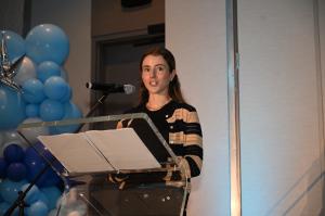 Keynote Speaker Cici Bellis, standing at the podium delivering her speech. She has long brown hair and is on a stage with a clear podium and blue balloons in the background.