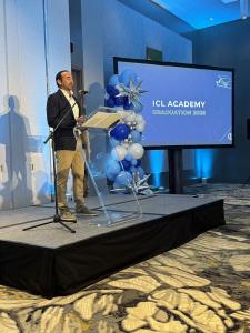 ICL Founder Kirk Spahn on stage delivering a speech at ICL Academy graduation. He has short brown hair and is wearing khaki pants with a white, button down shirt and a blue blazer. To his right side stands a large blue screen that says ICL Academy Graduat