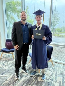 Two men standing next to each other smiling. On the left hand side is Dayton Hansen wearing a black suit and blue button down shirt, and to the right is an ICL graduate wearing a blue cap and gown, with khakis and a white shirt underneath, a lei around hi