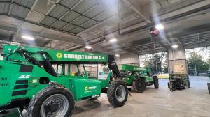 Interior view of a Sunbelt Rentals facility showing two green JLG telehandlers parked inside a maintenance bay. Overhead lighting and equipment, including hose reels and safety signage, are visible, with additional construction equipment seen outside thro