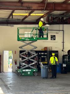 Two workers in safety gear perform an LED lighting upgrade inside a Sunbelt Rentals facility. One worker stands on a green scissor lift inspecting overhead fixtures, while the other stands below with a rolling bin. The maintenance area contains shelving w