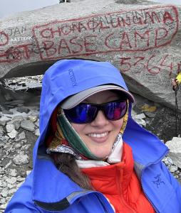 Woman wearing a orange and blue mountaineering jacket and reflective sunglasses smiles while standing infront of Mount Everest Base Camp Rock at 5,364 metres altitude on May 7, 2025.