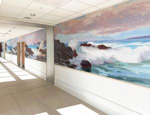 Hospital corridor featuring a mounted mural that spans the length of the wall. The painting depicts dramatic ocean waves crashing against rocky cliffs beneath a dynamic sky of purples, pinks, and blues. Sunlight from large windows creates geometric patter