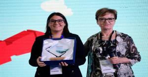 Two women at Maker Faire Rome 2024; one is holding a framed MakeITcircular award, while the other holds a microphone. They are standing in front of a colourful screen background.