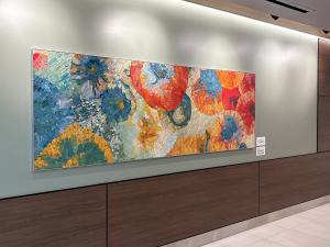 A colorful mosaic artwork mounted on a pale green hospital wall, featuring overlapping circular patterns in vibrant reds, oranges, blues, and yellows. The intricate tilework creates a dynamic, uplifting visual that enlivens the corridor space.
